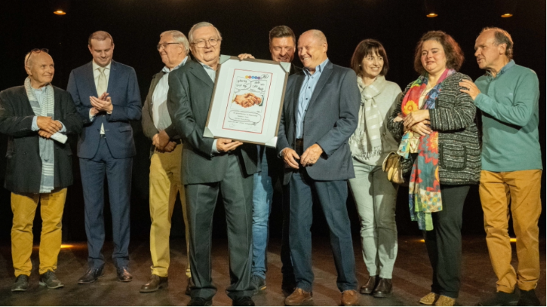 Voorzitter Guido Smets heeft net de ingekaderde tekening  int onvangst genomen. We zien v.l.n.r. Schepen Follens, Schepen Vancraeynest, Freddy Nachtegael, voozitter Guido Smets, Burgemeester Koen Metsu, Edgar De Keulenaer, ex leerkracht Maria Baldor en huidig leerkracht Spaans Carolina Merino en Schepen Muyters tevens voorzitter van de gemeenteraad.Schepen Brigitte Vermeulen-Goris en Koen Michiels vielen net naast de foto.